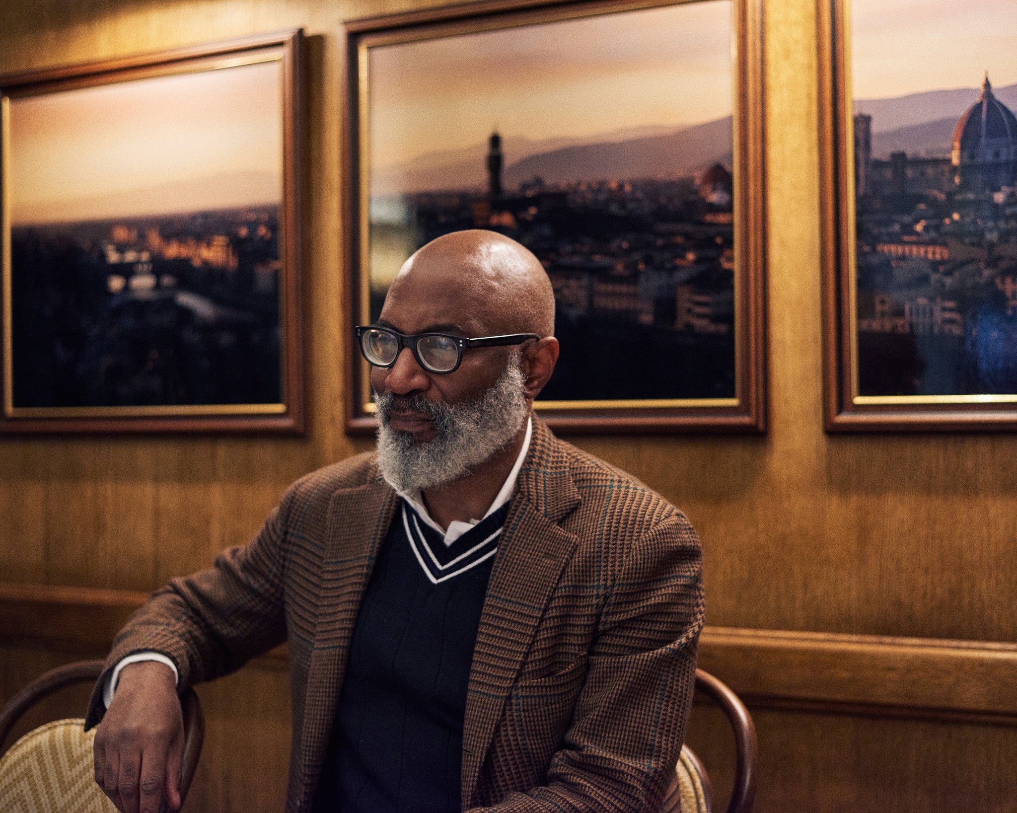 Jason Jules sitting in a brown room against a triptych of a skyline looking away from the camera, previously a designer and stylist, he worked closely in fashion circles in the 80s. Read the interview at A Collected Man London.