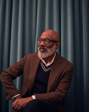 Jason Jules in a brown suit and white collared shirt against the background of a blue curtain, speaking about Ivy Style, African American fashion, jazz, photography, and more. Read the interview now at A Collected Man London.