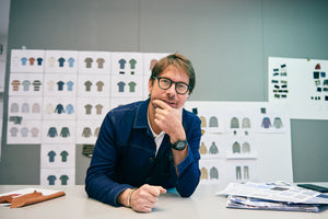 Oliver Spencer menswear designer standing in front of a green background covered in shirt designs and other trouser designs interviewed by A Collected Man London