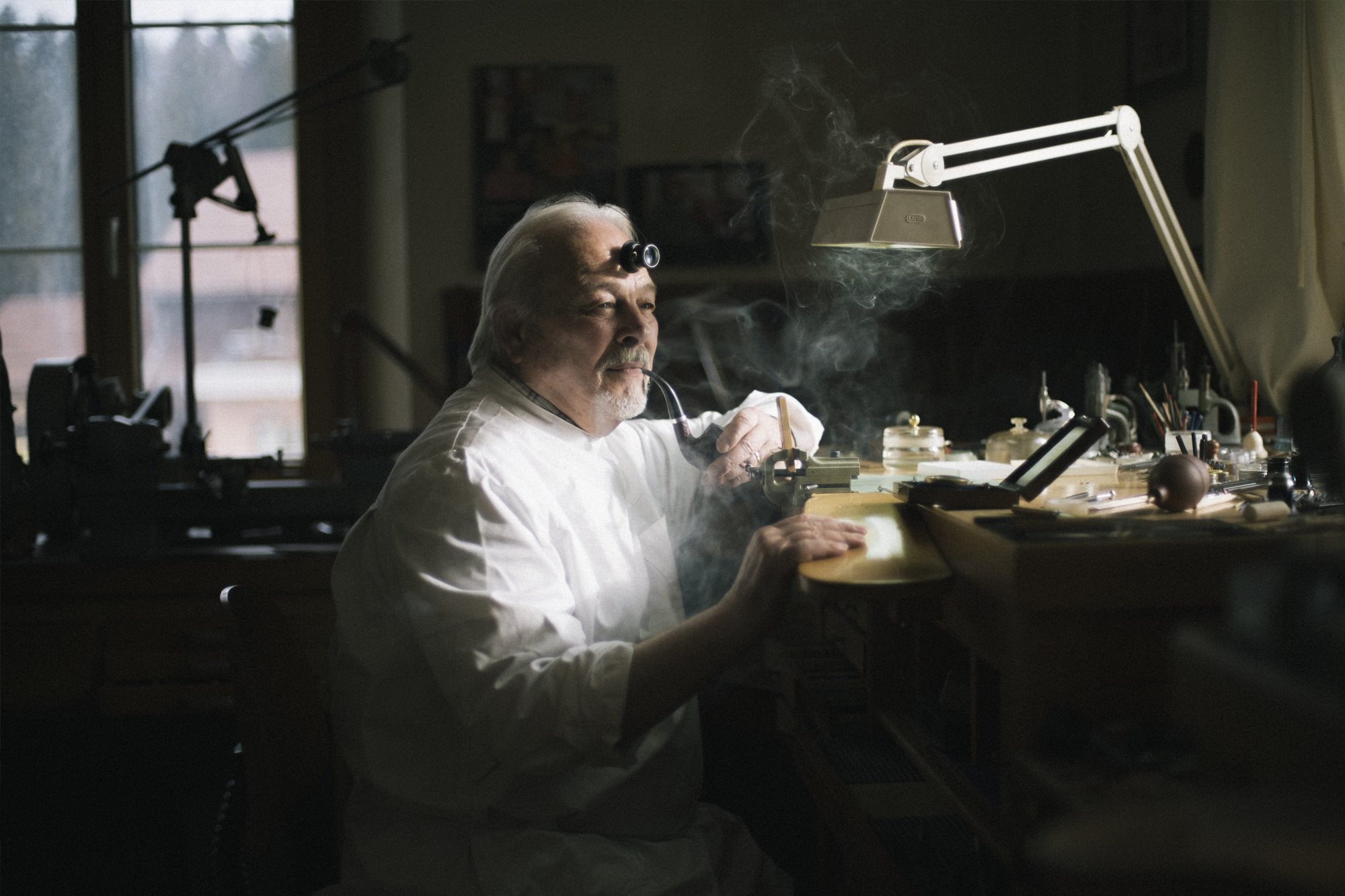 Philippe Dufour an older man with white hair wearing a white shift and surrounded by watchmaking tools in his office
